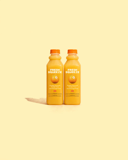 Two plastic bottles filled with orange juice and topped with orange caps, standing side by side on a pale yellow background.