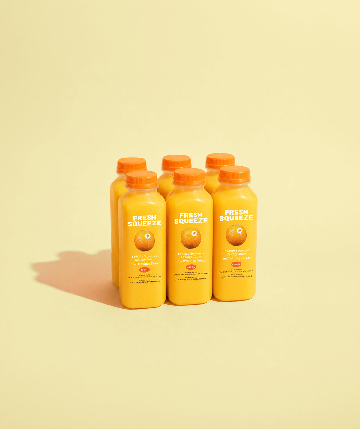 Six small square bottles filled with bright orange juice, arranged in two rows on a pale cream background with a soft shadow.